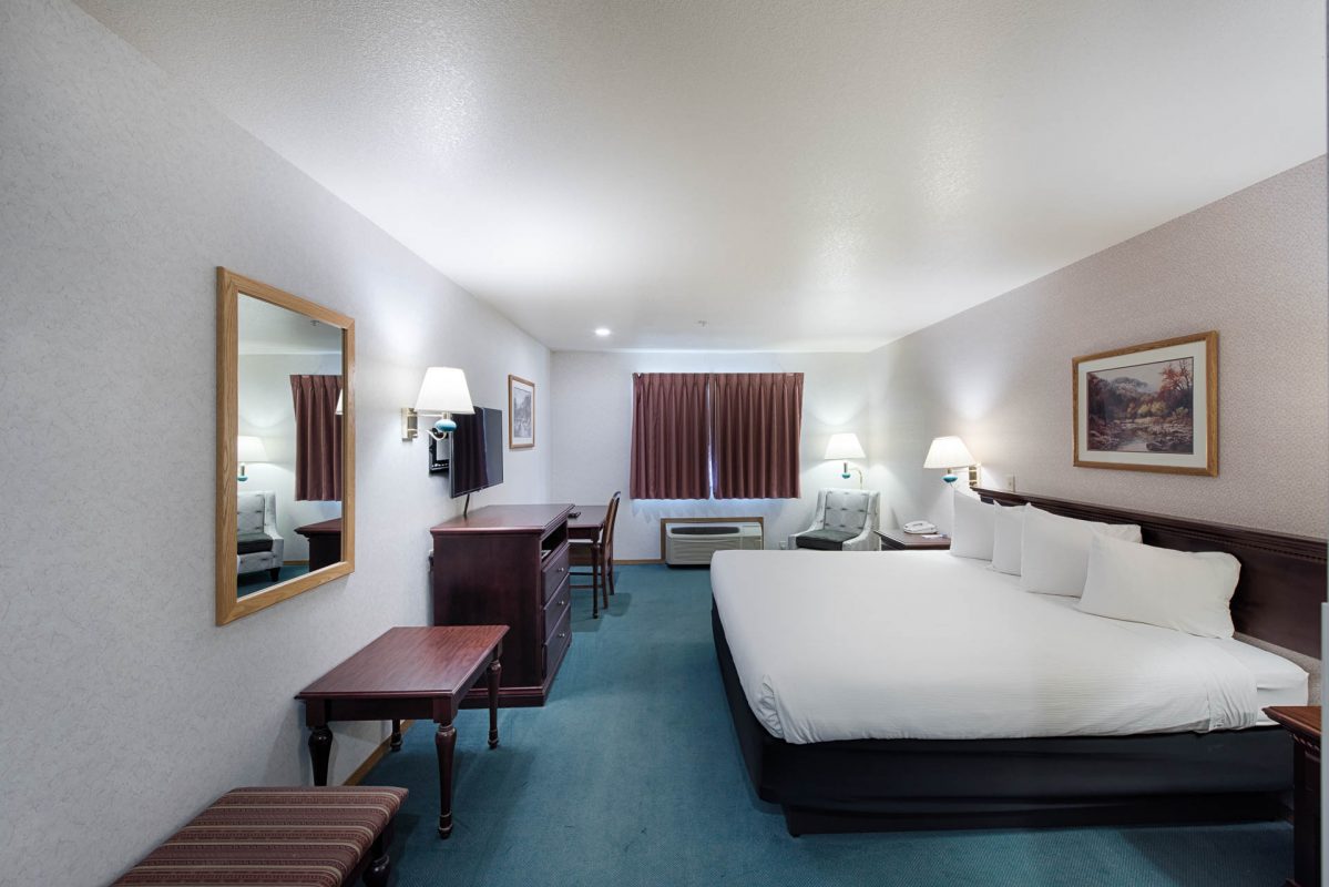 A wide-angle view of a hotel room with a large bed, desk, mirror, TV, and window.